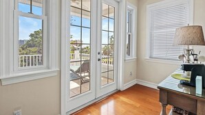 Una habitación con un gran ventanal corredera que da a un balcón, una mesita con una lámpara y una ventana con persianas.