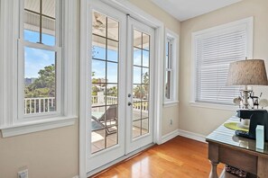 Una habitación con un gran ventanal corredera que da a un balcón, una mesita con una lámpara y una ventana con persianas.