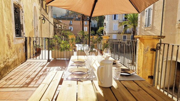 An outdoor patio with a wooden table set for dining, surrounded by a metal railing and potted plants.