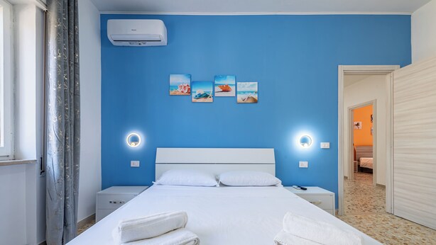 Een hotelkamer met een blauwe muur, een bed met witte lakens, een nachtkastje, een wandairconditioner en een deur naar een andere kamer.