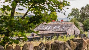A rustic stone barn with a sloped roof and multiple skylights, surrounded by a wooden fence and lush greenery.