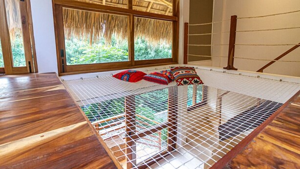 A room with a wooden floor, a bed with a mosquito net, and a large window with a view of greenery. - 2 Room Loft w/ patio, parking, kitchen (Mazunte)