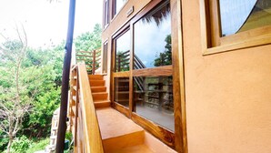 Un escalier en bois menant à un balcon doté d’une porte vitrée, le tout entouré de verdure.
