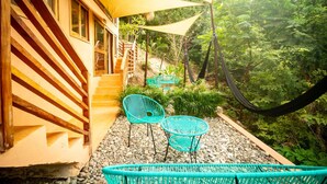 Un escalier en bois mène à un bâtiment à la façade jaune, avec un espace extérieur aménagé de chaises turquoise et d’un hamac.