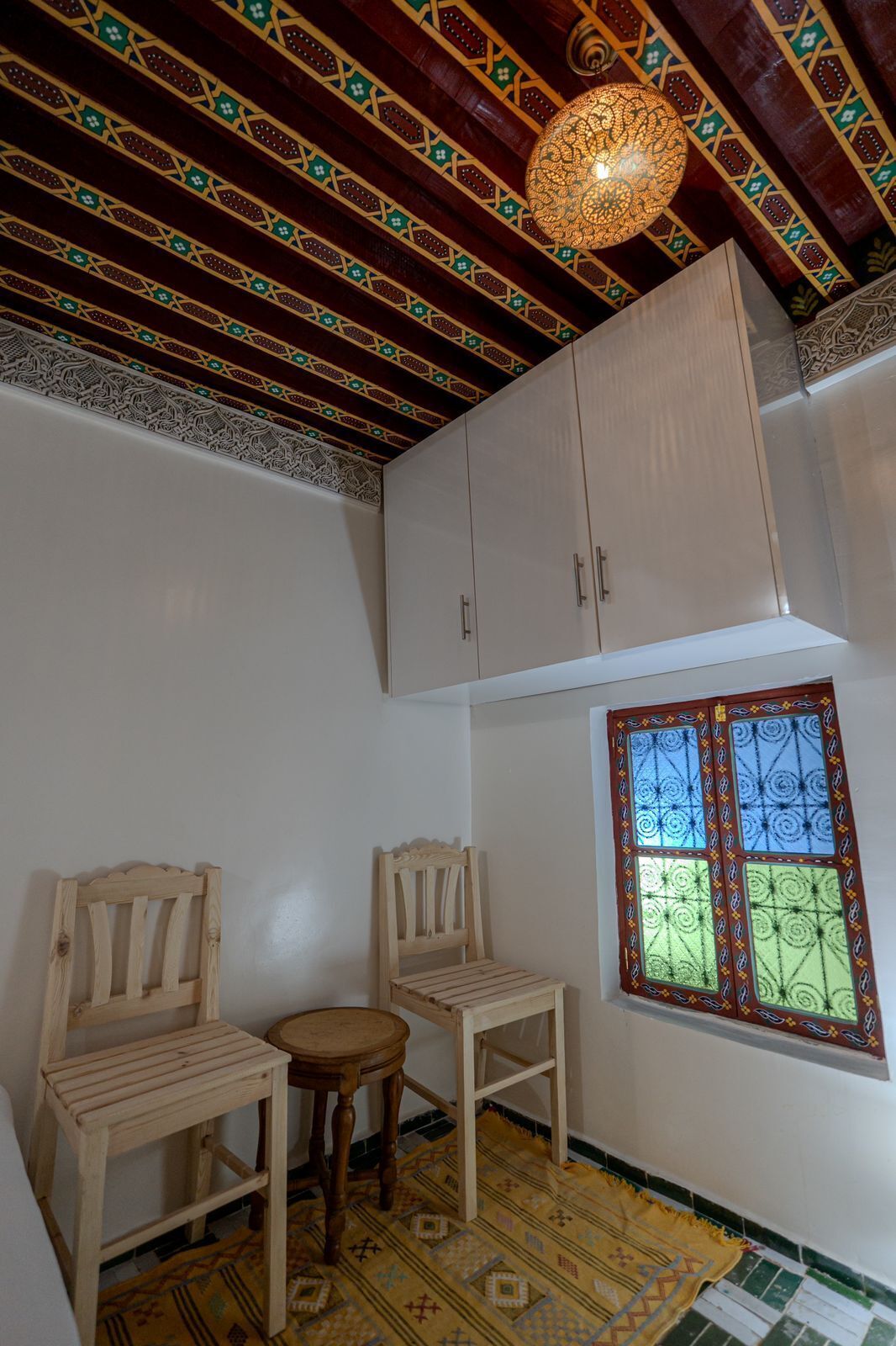 Une pièce avec un plafond orné de motifs, une fenêtre dotée d’un cadre décoratif et deux chaises en bois.