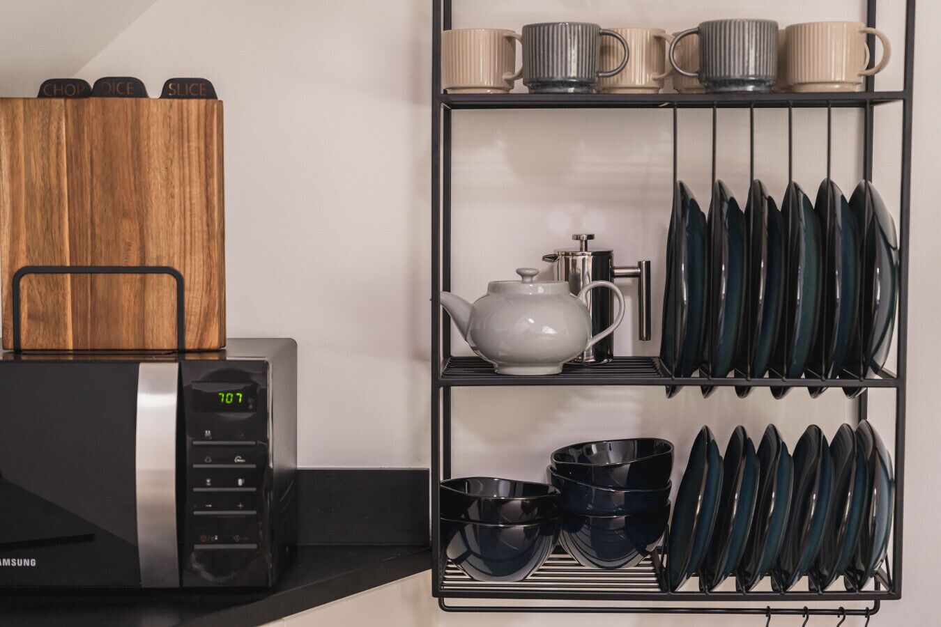 A kitchen area with a microwave, a wooden cutting board, a tea pot, and a rack holding plates, bowls, and cups.