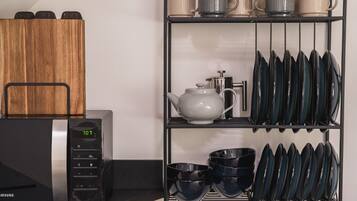 A kitchen area with a microwave, a wooden cutting board, a tea pot, and a rack holding plates, bowls, and cups.