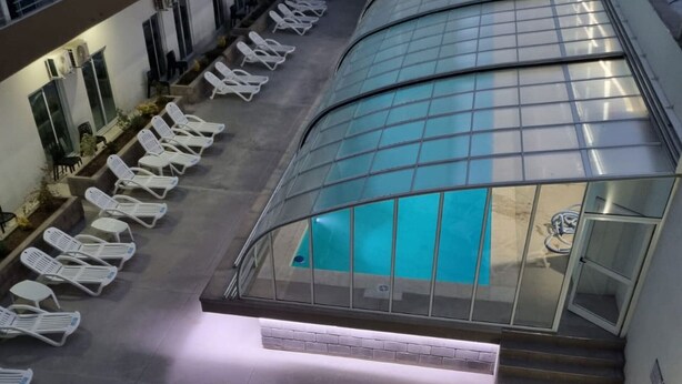 Un área de piscina interior moderna con cerramiento de vidrio, múltiples sillas de descanso y una escalera que lleva a un nivel superior.