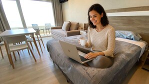 Una persona sentada en una cama con una computadora portátil y una taza, en una habitación con comedor y vistas al exterior.