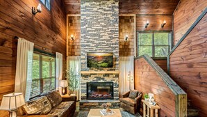 A cozy living room with a stone fireplace, wooden walls, a comfortable sofa, and a television. - Ultimate Mountain Getaway Pickleball & Basketball Courts Games Hot Tub Sleeps 20 (Sevierville)