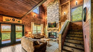 A log cabin interior with a stone fireplace, a sofa, and a staircase. - Ultimate Mountain Getaway Pickleball & Basketball Courts Games Hot Tub Sleeps 20 (Sevierville)