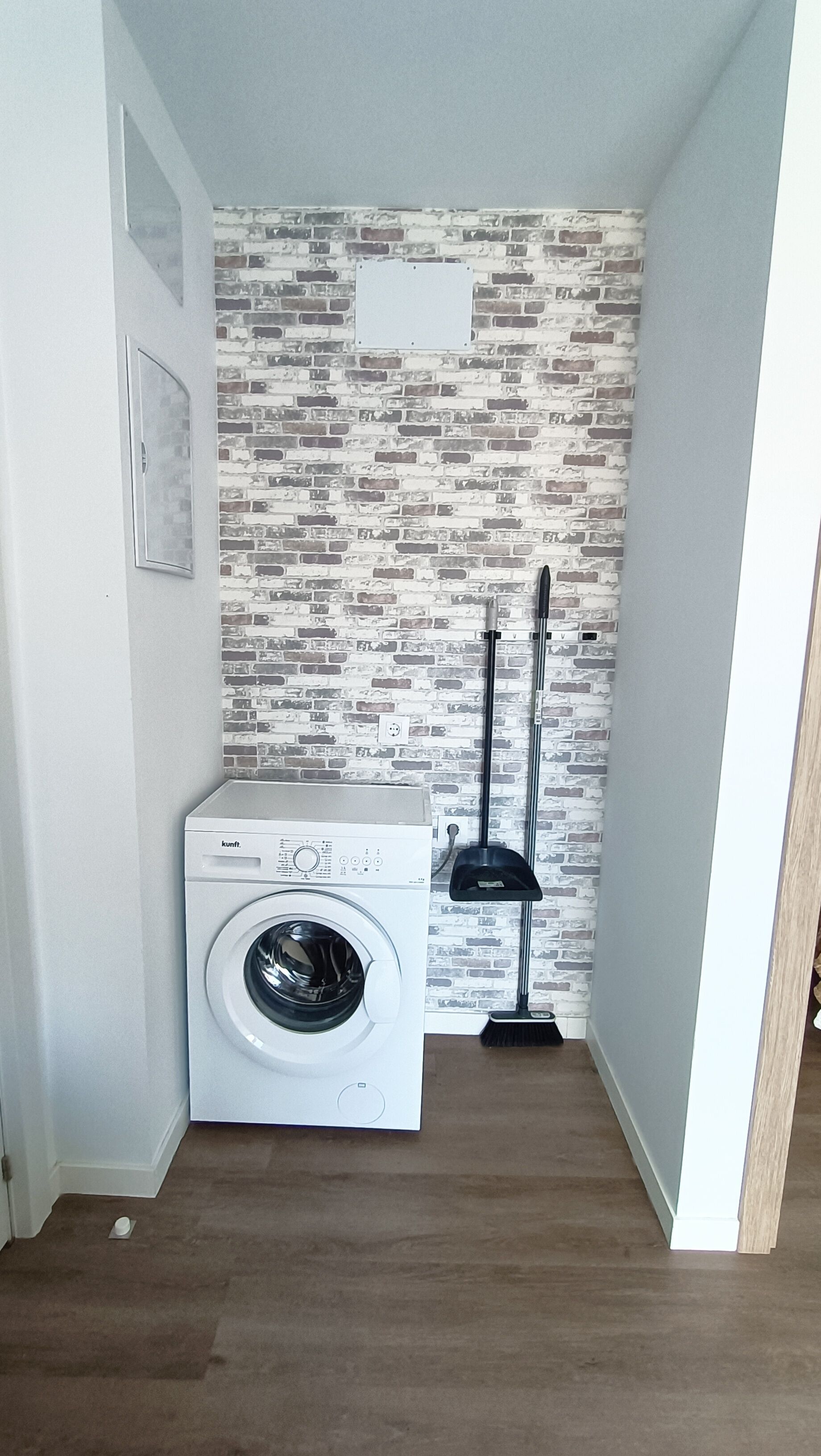 Un cuarto de lavado con una lavadora blanca, un escoba y un pared con diseño de ladrillo.