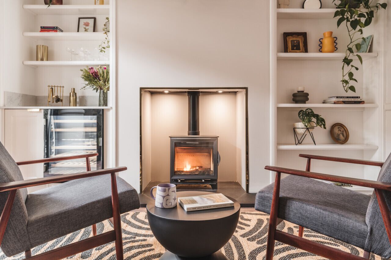 A cozy living room with a fireplace, two armchairs, a coffee table, and shelves with books and decorative items.