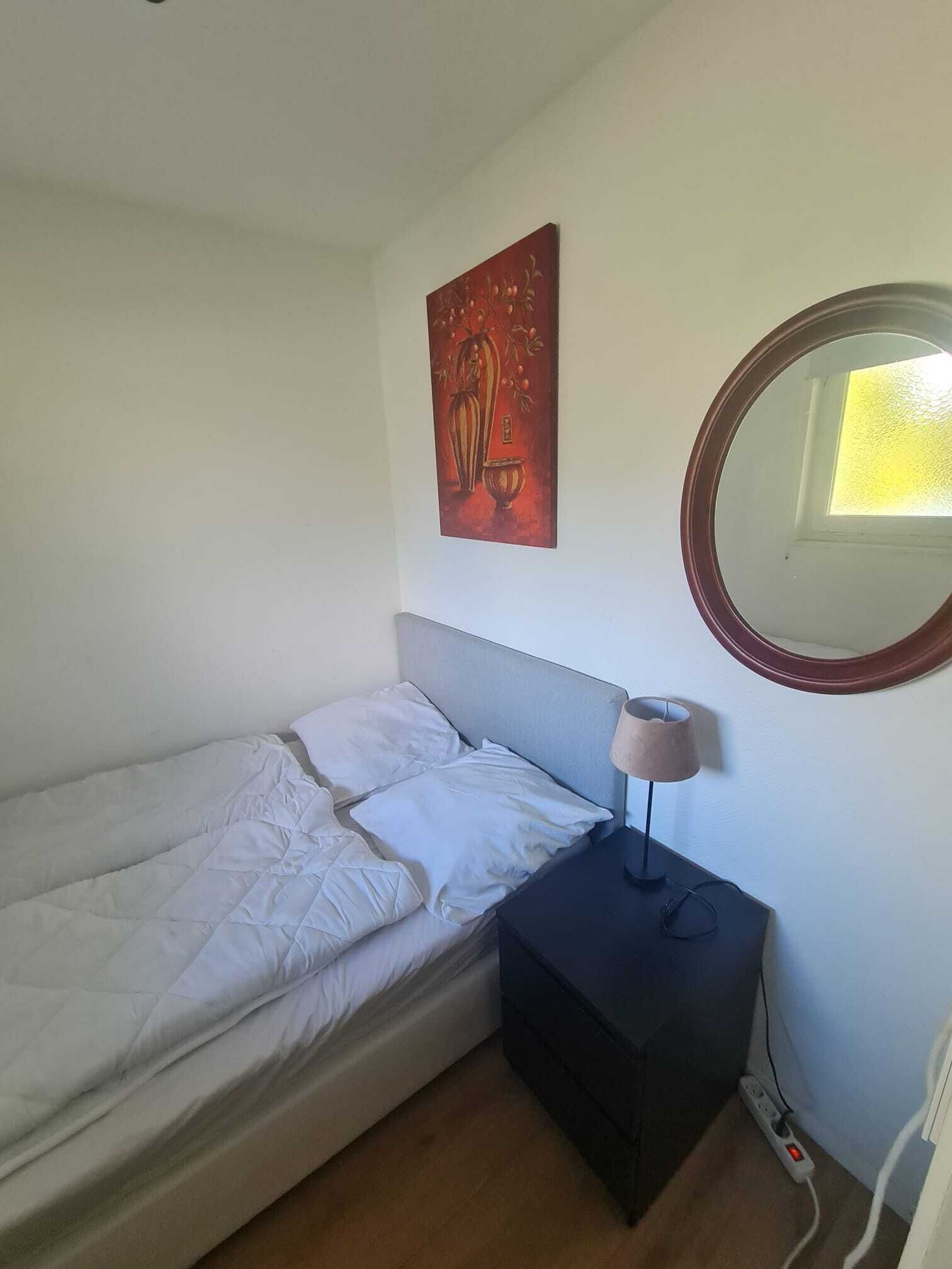 Una cama individual con ropa de cama blanca, una mesita de noche con lámpara, una regleta y un espejo fijado a la pared.