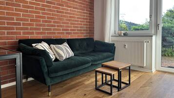 Una habitación con un sofá verde, una mesa de centro de madera, una pared de ladrillo y una ventana con vistas a la vegetación.