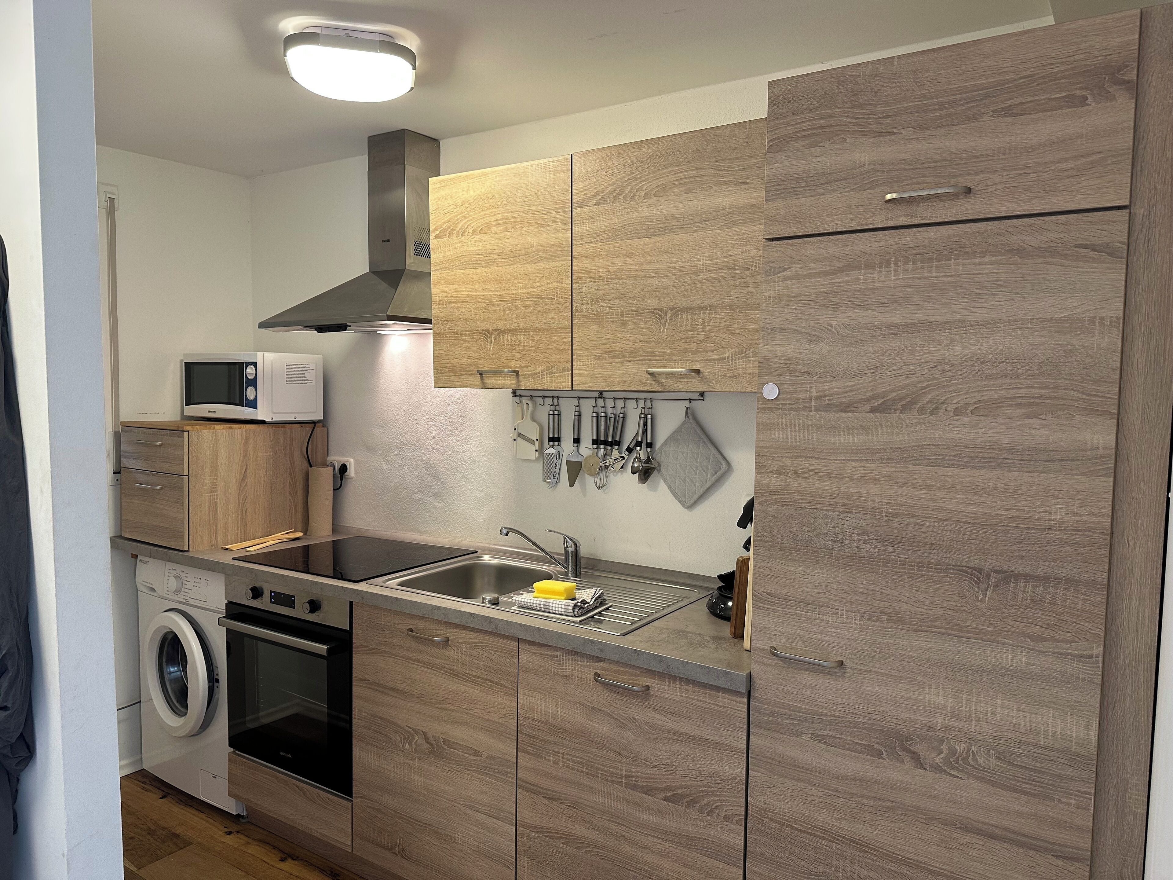 Een kleine keuken met houten kastjes, een magnetron, een wasmachine en een spoelbak.