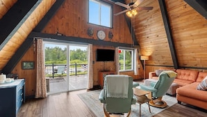 Ein gemütliches Loft-Zimmer mit Holzbalkendecke, Deckenventilator, Flachbildfernseher, einem grünen Sessel, einem orangefarbenen Sofa und einem Esstisch.