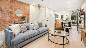 A modern living room with a brick wall, a round mirror, a grey sofa with patterned cushions, a coffee table, and a staircase leading to an upper floor.