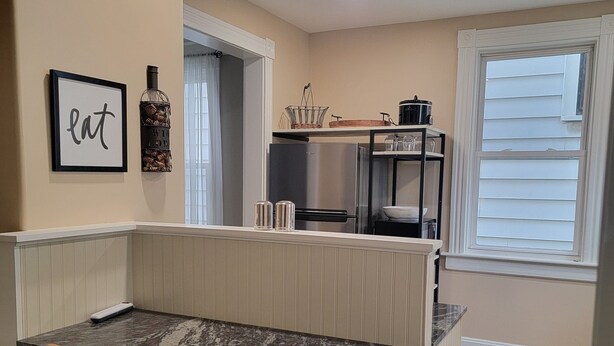 A compact kitchen with a built-in island, two foldable chairs, a refrigerator, and a window with blinds. - 1895 meets 2025 Townhouse in Urban Hamilton (Hamilton)
