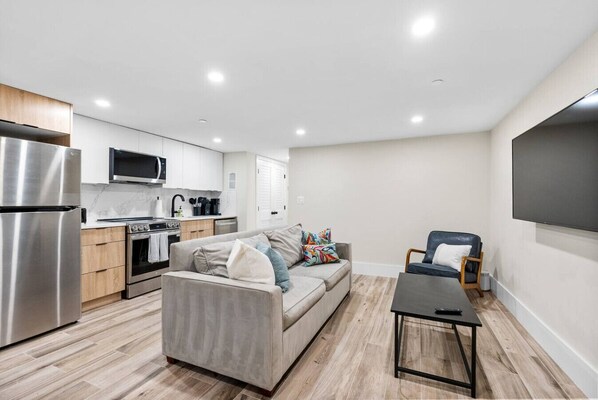 Een modern appartement met een kitchenette, een grijze bank, een kleine tafel en een aan de muur bevestigde televisie.
