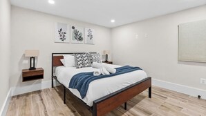 Une chambre moderne avec un lit en bois, une literie blanche et une couverture bleue. On y trouve deux tables de chevet avec des lampes, et une œuvre d’art accrochée au mur au-dessus du lit.