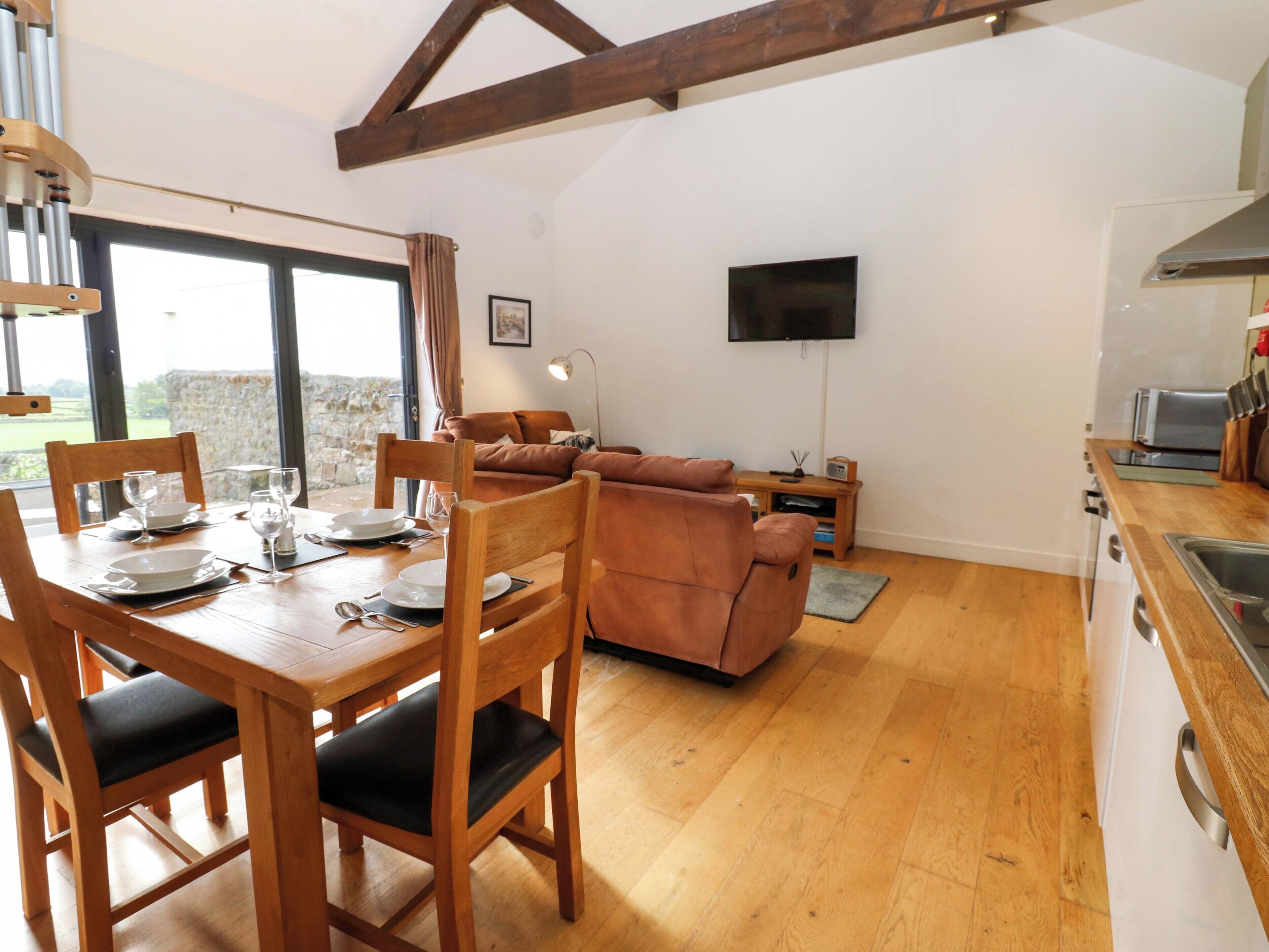 A dining area with a wooden table and chairs, a sofa, a TV mounted on the wall, and a kitchen area with a sink and stove.