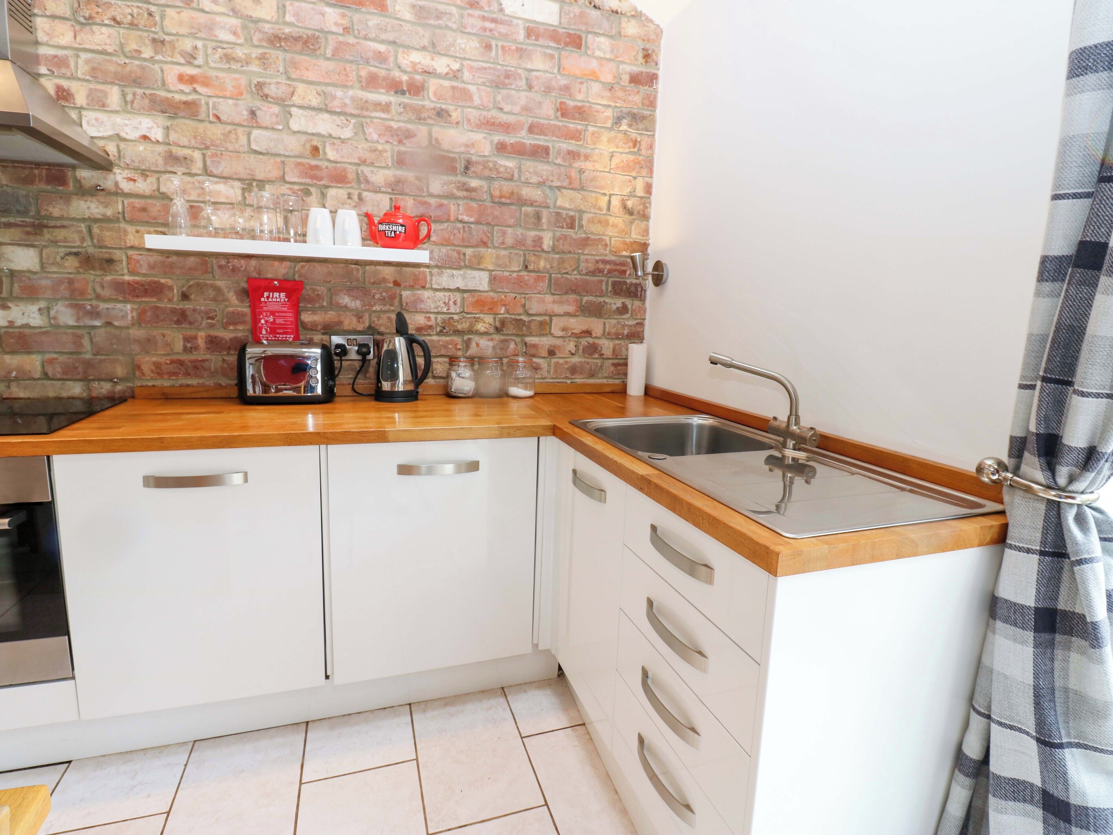 A kitchen with a brick wall, white cabinets, a wooden countertop, a toaster, a kettle, and a sink.