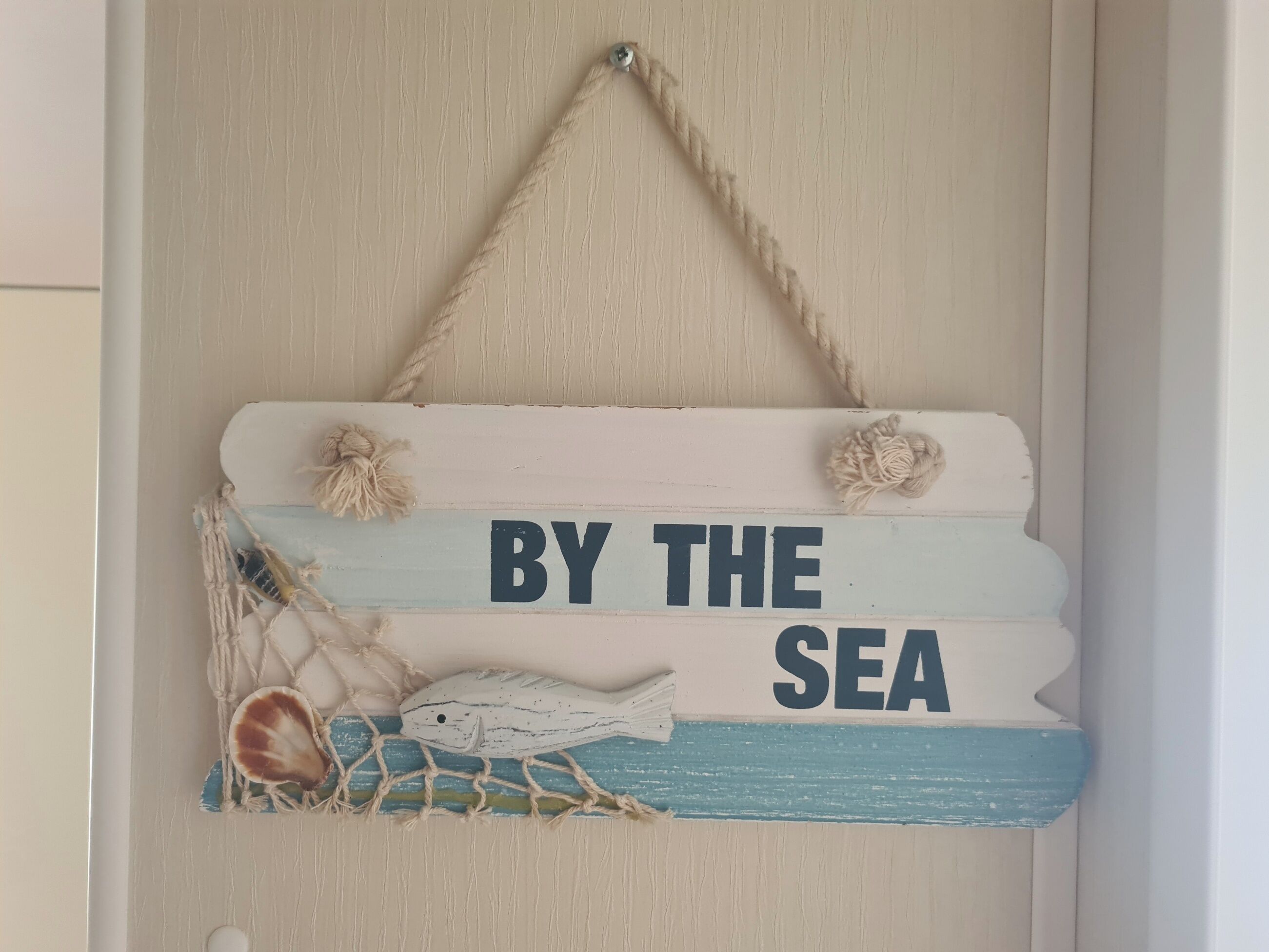 A decorative sign with the text "BY THE SEA" and nautical elements like a fish and net.