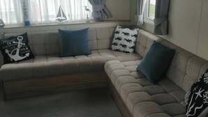 Una sala acogedora con un sofá seccional color beige, dos almohadas azules y una almohada estampada en blanco y negro. Se observa un reloj de pared con temática náutica y un cuadro de una escena costera.