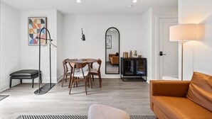 Een moderne woonkamer met een ronde houten tafel, een bank, een vloerlamp en een spiegel die een eethoek weerspiegelt.
