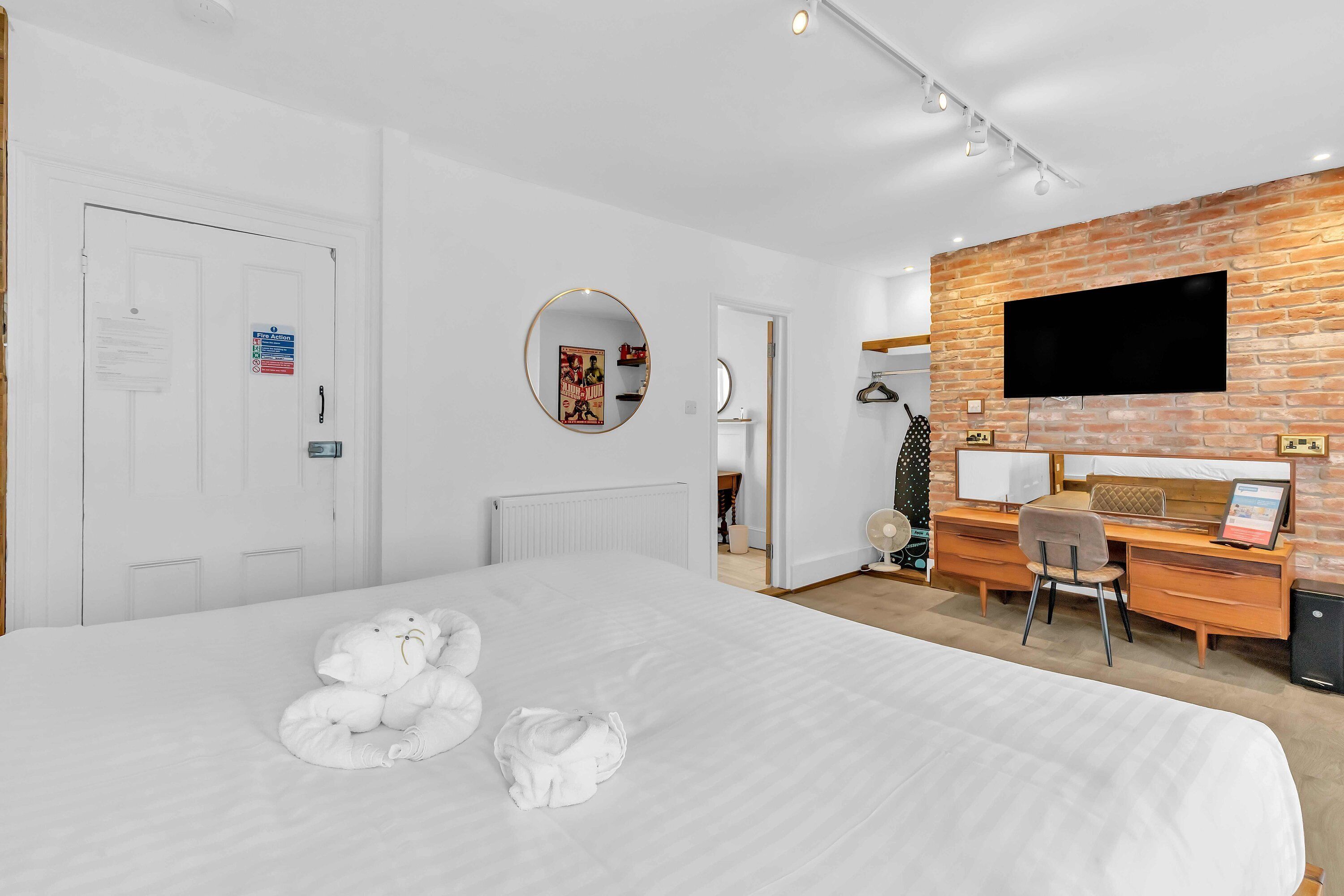 A hotel room with a large bed, a desk with a computer, a TV mounted on a brick wall, and a white door with a fire action sign.
