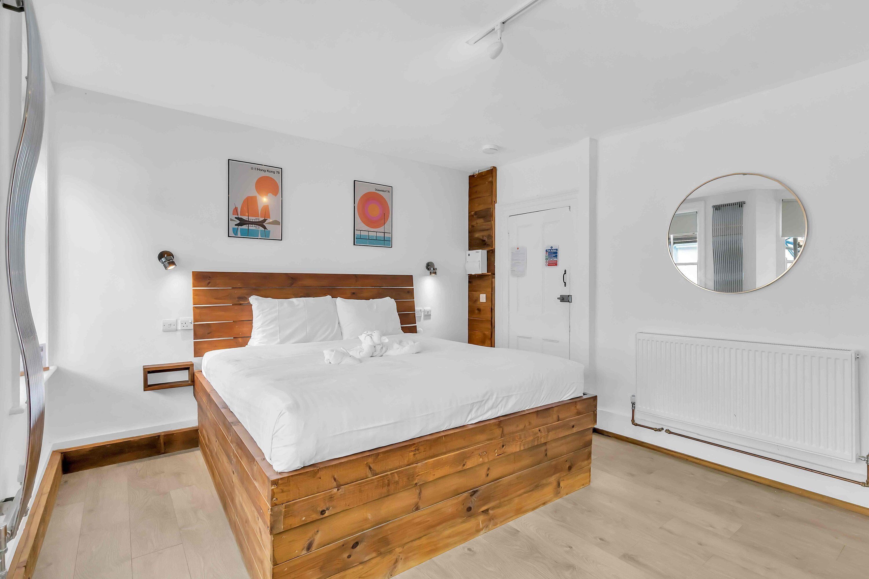 A bedroom with a wooden bed frame, white bedding, a round mirror, and two wall-mounted light fixtures.