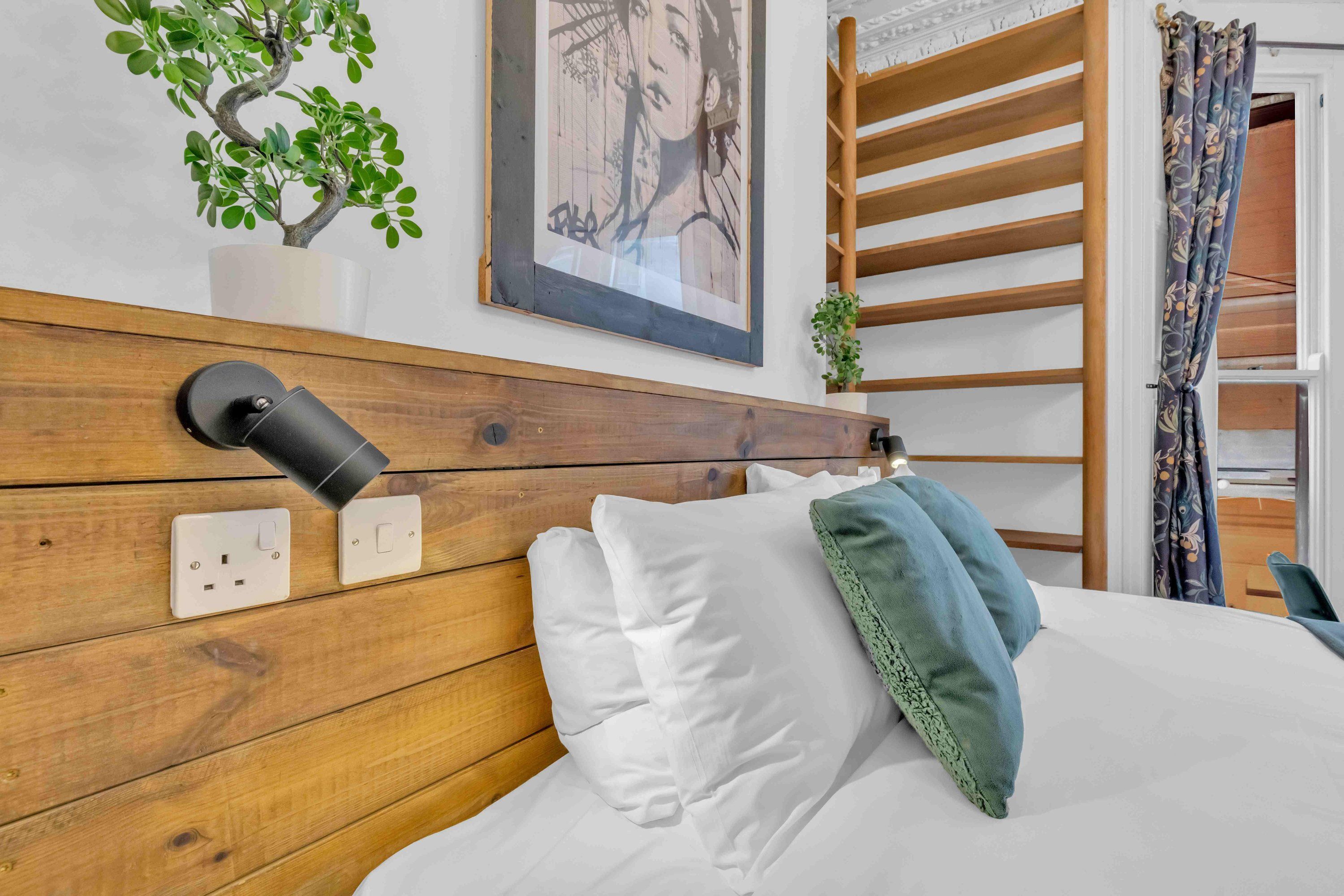 Een slaapkamer met een houten hoofdeinde, een nachtkastje met een lamp, een stopcontact en een ingelijst beeld aan de muur.