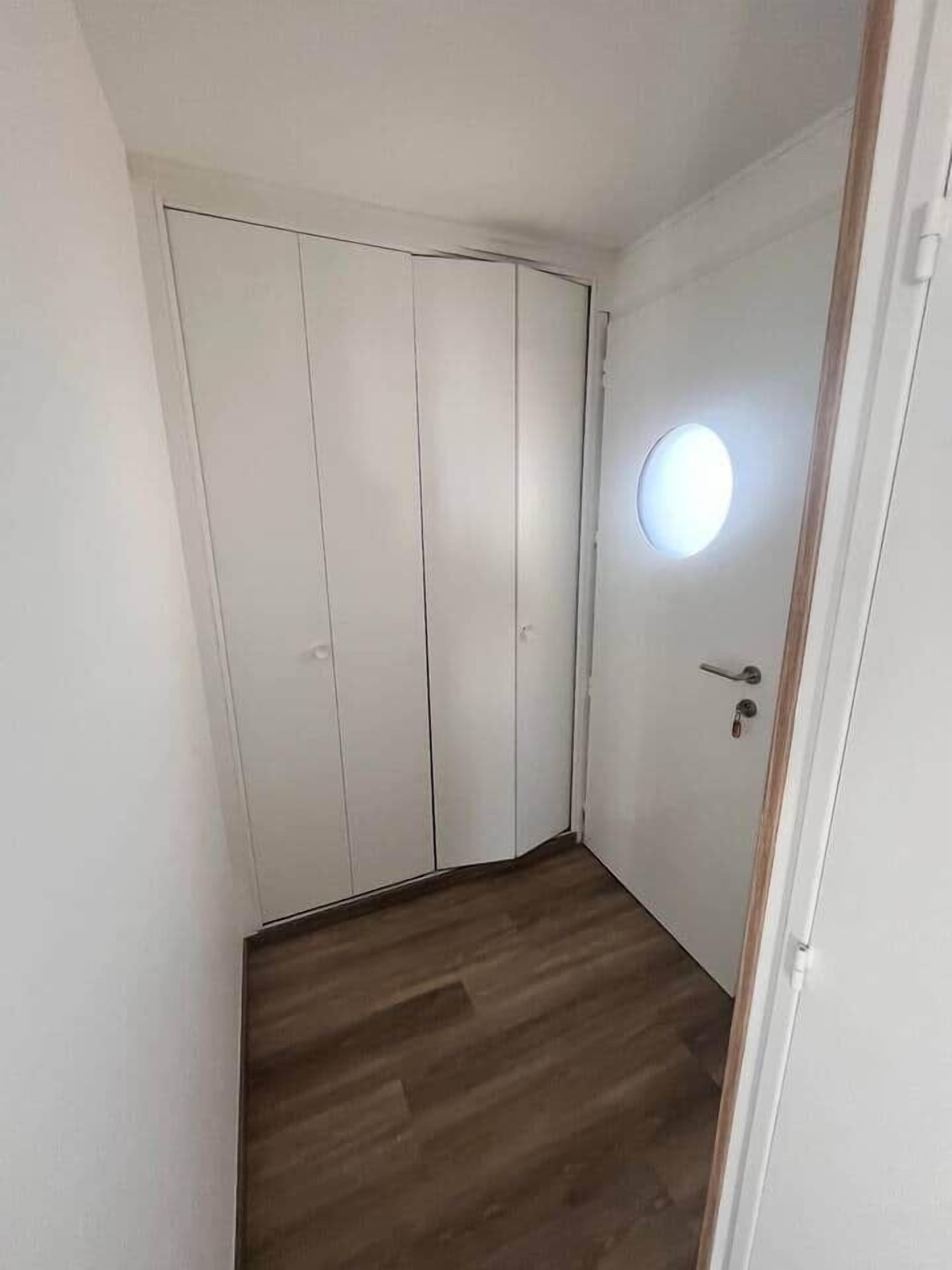 Een smalle gang met een houten vloer en een witte deur met een rond raam.