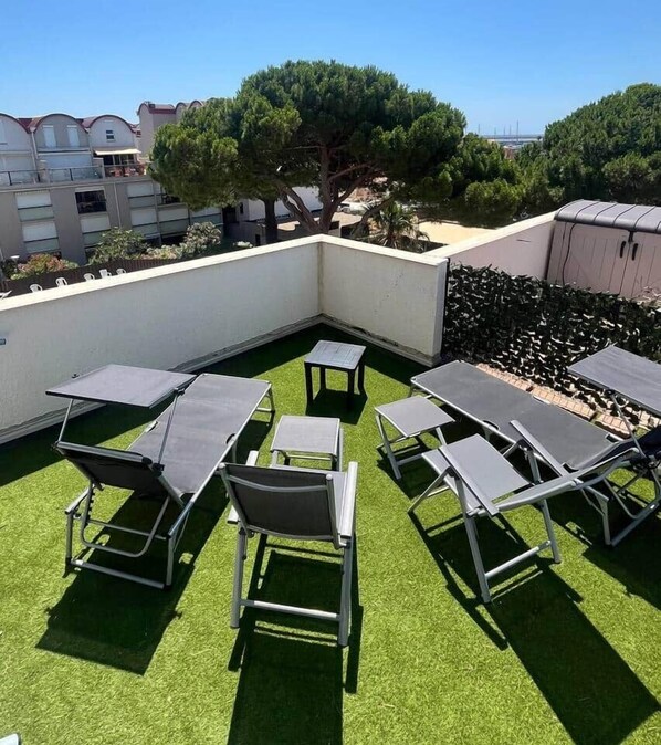 An outdoor seating area with tables and chairs on artificial grass, surrounded by a white wall and a hedge. - Air-Conditioned Two-Bedroom Apartment / Large Terrace (Gruissan)