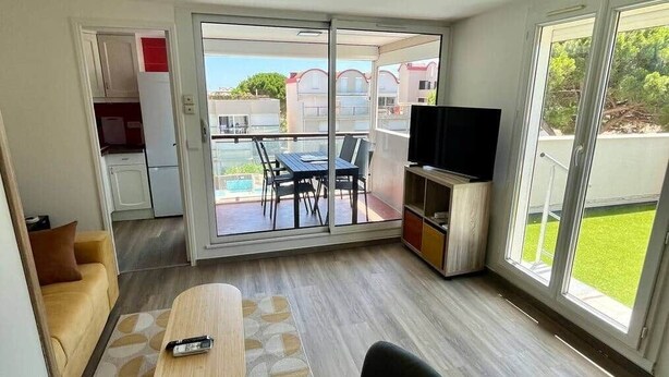 Een moderne woonkamer met een bank, een salontafel en een televisie. Er is een glazen schuifdeur die leidt naar een buitenruimte met een zwembad en uitzicht op gebouwen.