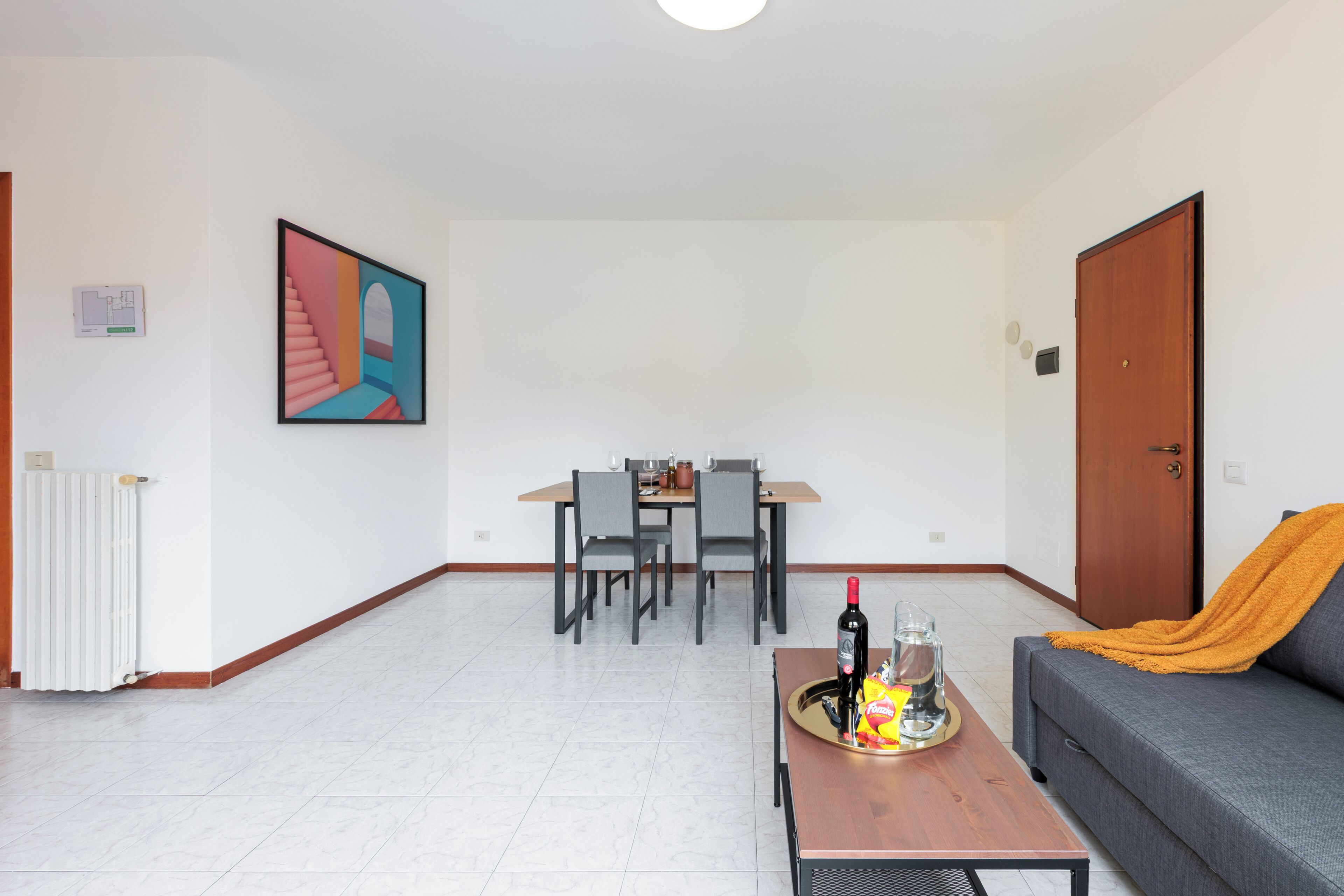 Ein Wohnzimmer mit einer Couch, einem Couchtisch mit einer Flasche und Snacks, einem Essbereich mit Stühlen und einem Tisch sowie einer Tür.