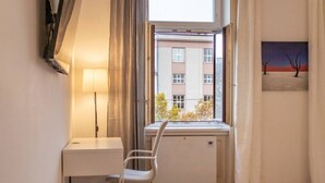 Een kamer met een raam, een radiator, een bureau, een stoel en een schilderij aan de muur.