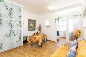 Un comedor con una mesa preparada para cuatro personas, un sofá con cojines, un cuadro en la pared y una puerta que da a otra habitación.