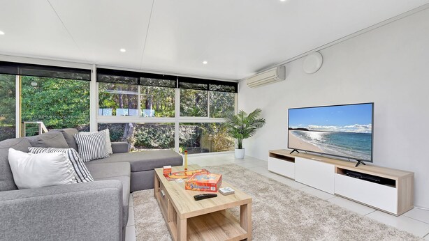 A modern living room with a large TV, grey sofa, and a view of the outdoors through large windows. - Oasis at Salamander Bay (Salamander Bay)