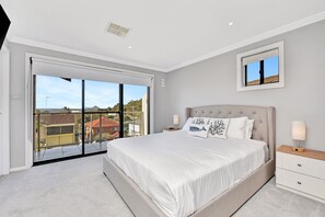 A bedroom with a large bed, a nightstand, a bedside lamp, a window with a view of buildings, and a ceiling vent.