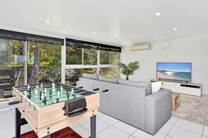 A modern living room with a foosball table, a grey sofa, a television, and large windows offering a view of greenery.