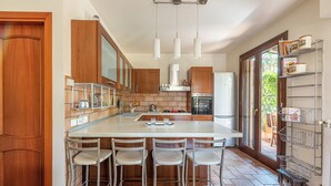 A modern kitchen with wooden cabinets, a large island with bar stools, a built-in oven, and a window overlooking greenery.