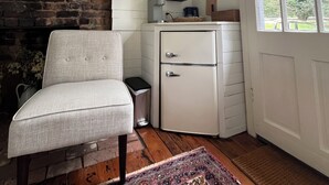 A room with a white chair, a small white refrigerator, a fireplace, a window with a view of greenery, and a patterned rug. - Cozy Cottage Perfect for a Charming Charlottesville (Charlottesville)