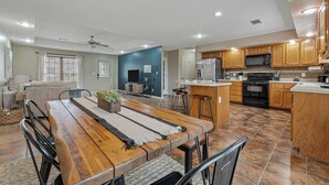 Uma sala de estar ampla com uma mesa de jantar de madeira, um sofá e uma televisão. A cozinha possui armários de madeira e um micro-ondas.