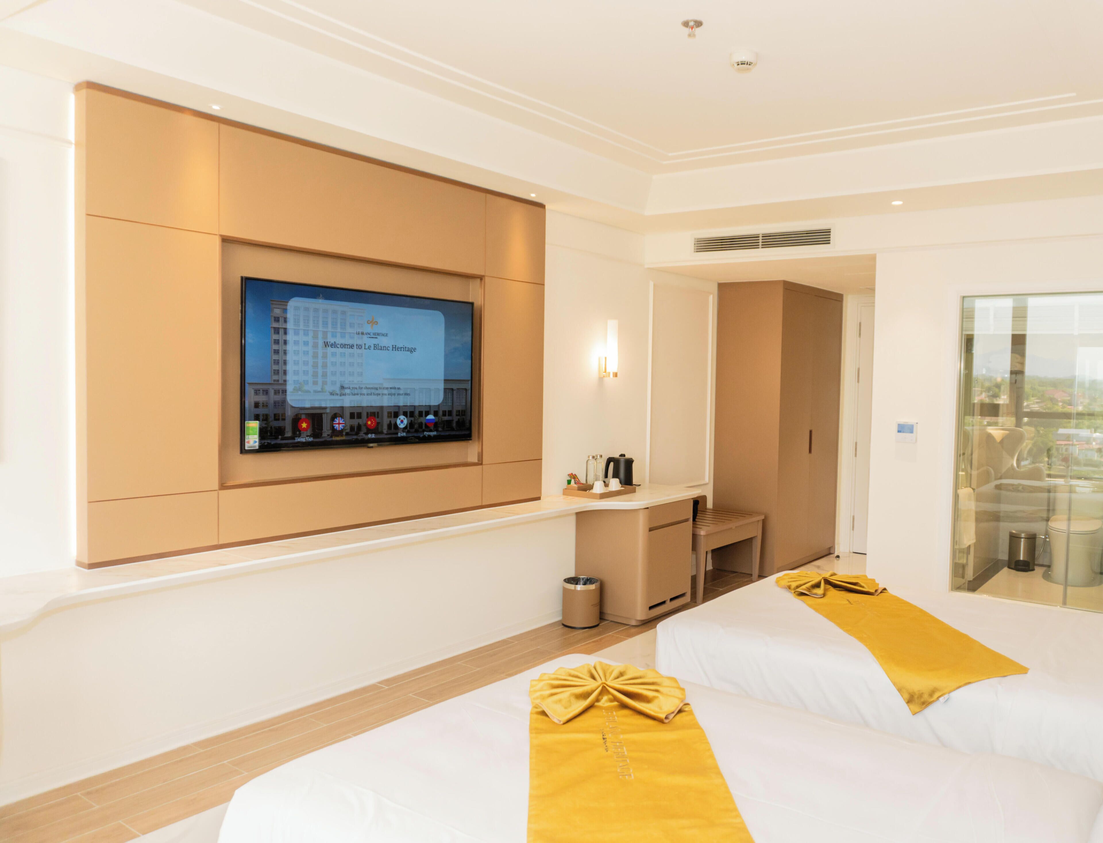 Ett hotellrum med en tv som är monterad på väggen, ett skrivbord med en stol, en säng med ett gult överkast och utsikt genom en glasdörr.