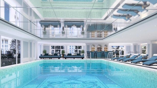 A modern indoor pool with clear glass walls, lounge chairs, and a bar area.