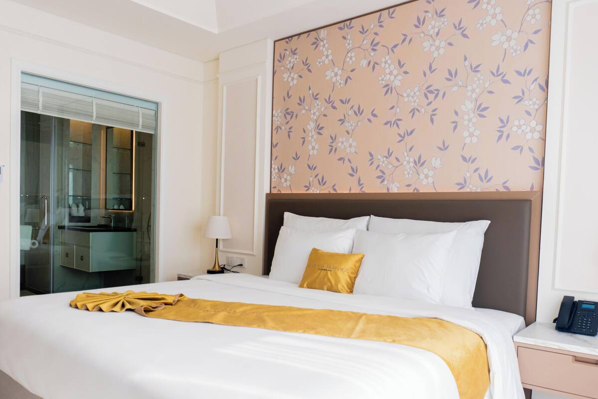 Habitación de hotel con cama, mesita de noche, teléfono, lámpara, espejo y baño con puerta de cristal.