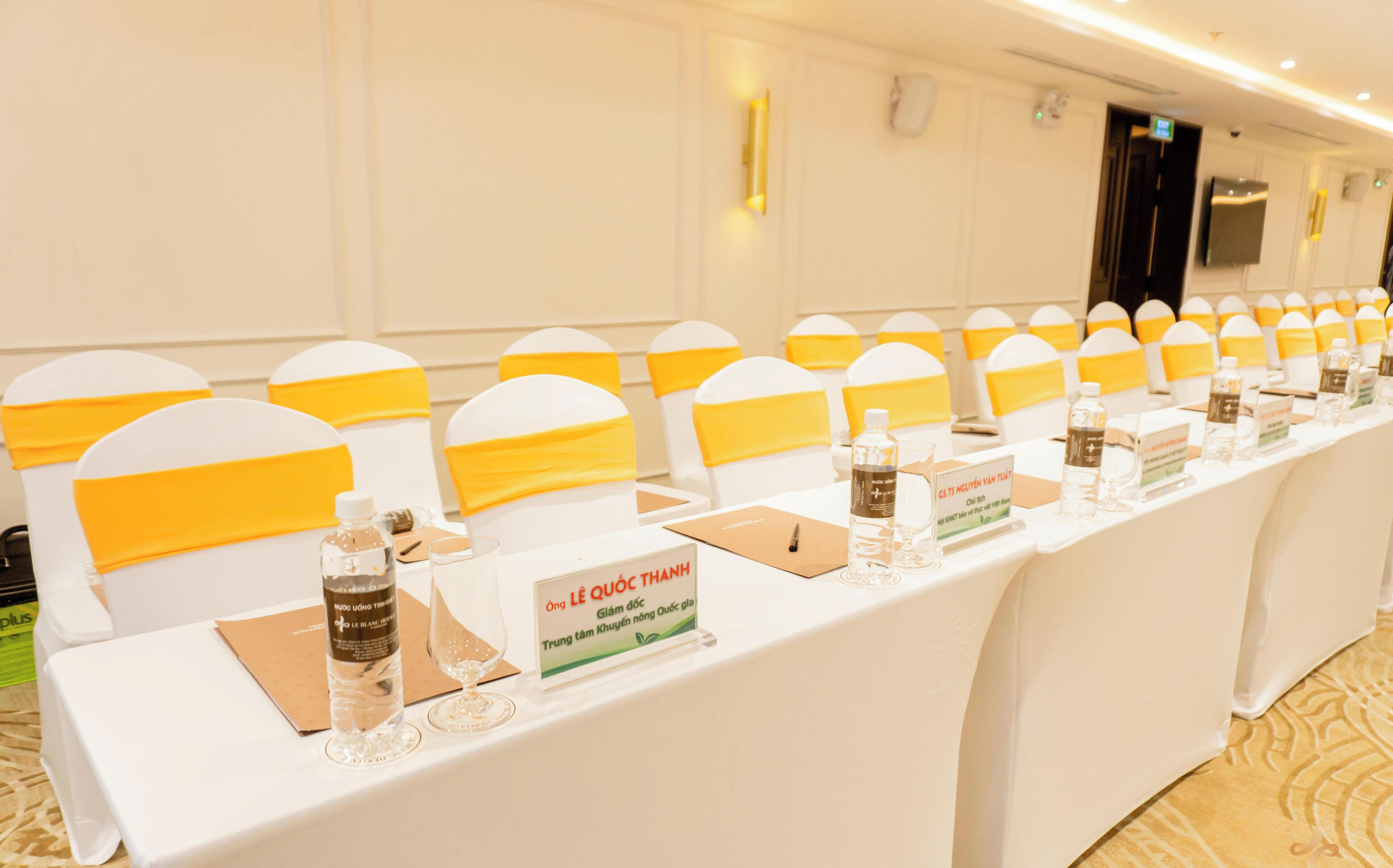 Un salón de conferencias con filas de sillas blancas, cada una cubierta con plástico de polietileno. Las mesas están preparadas con botellas de agua, vasos y tarjetas de identificación.