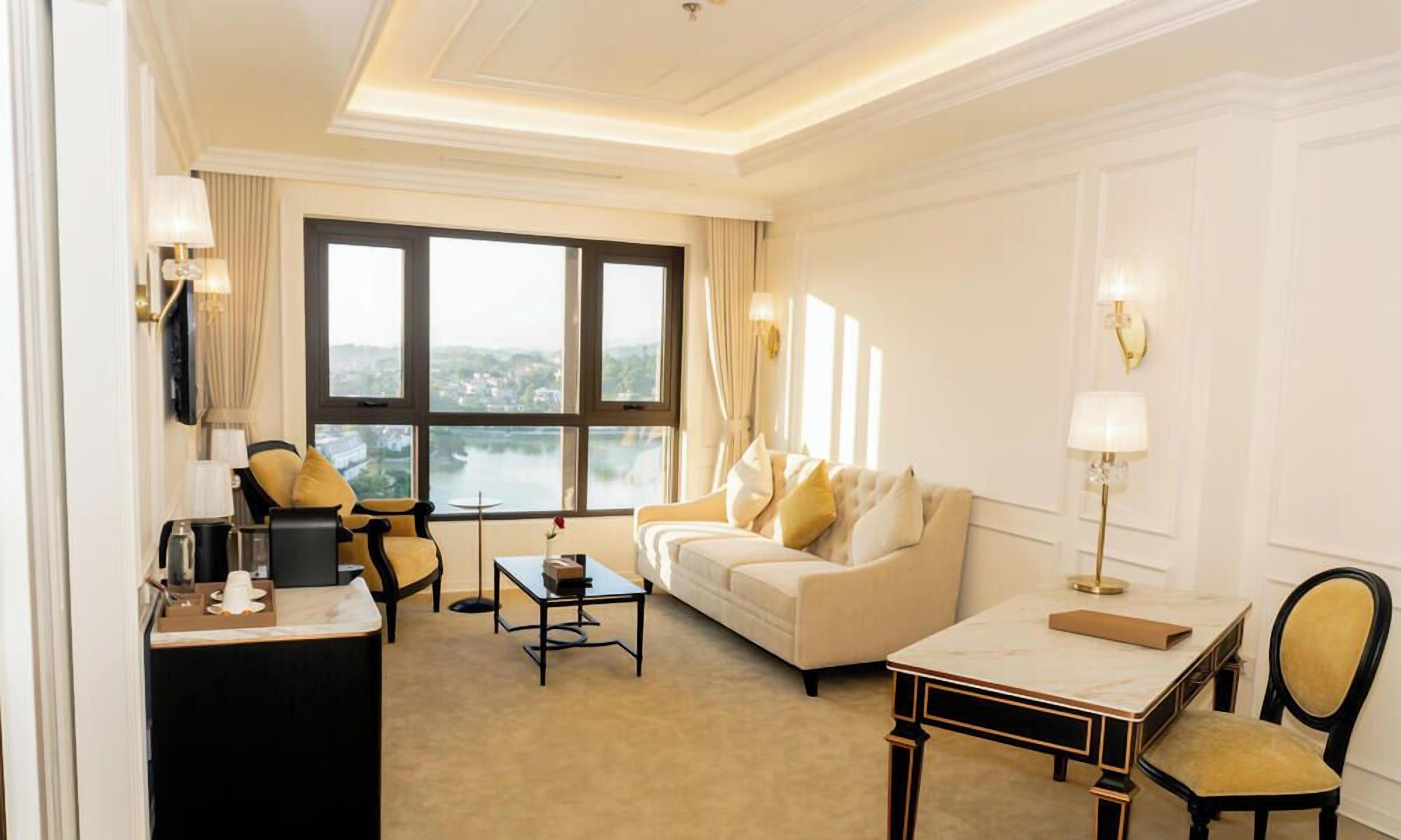 A hotel room with a large window, a sofa, a coffee table, a desk, and a chair.
